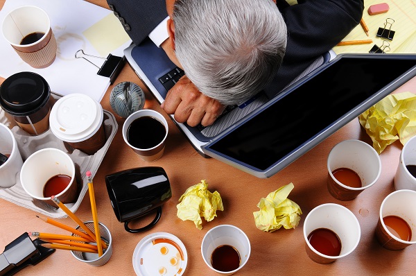 Closeup view of a very cluttered businessman's desk. Overhead view with man's head on laptop keyboard and scattered coffee cups and office supplies. Horizontal format.
